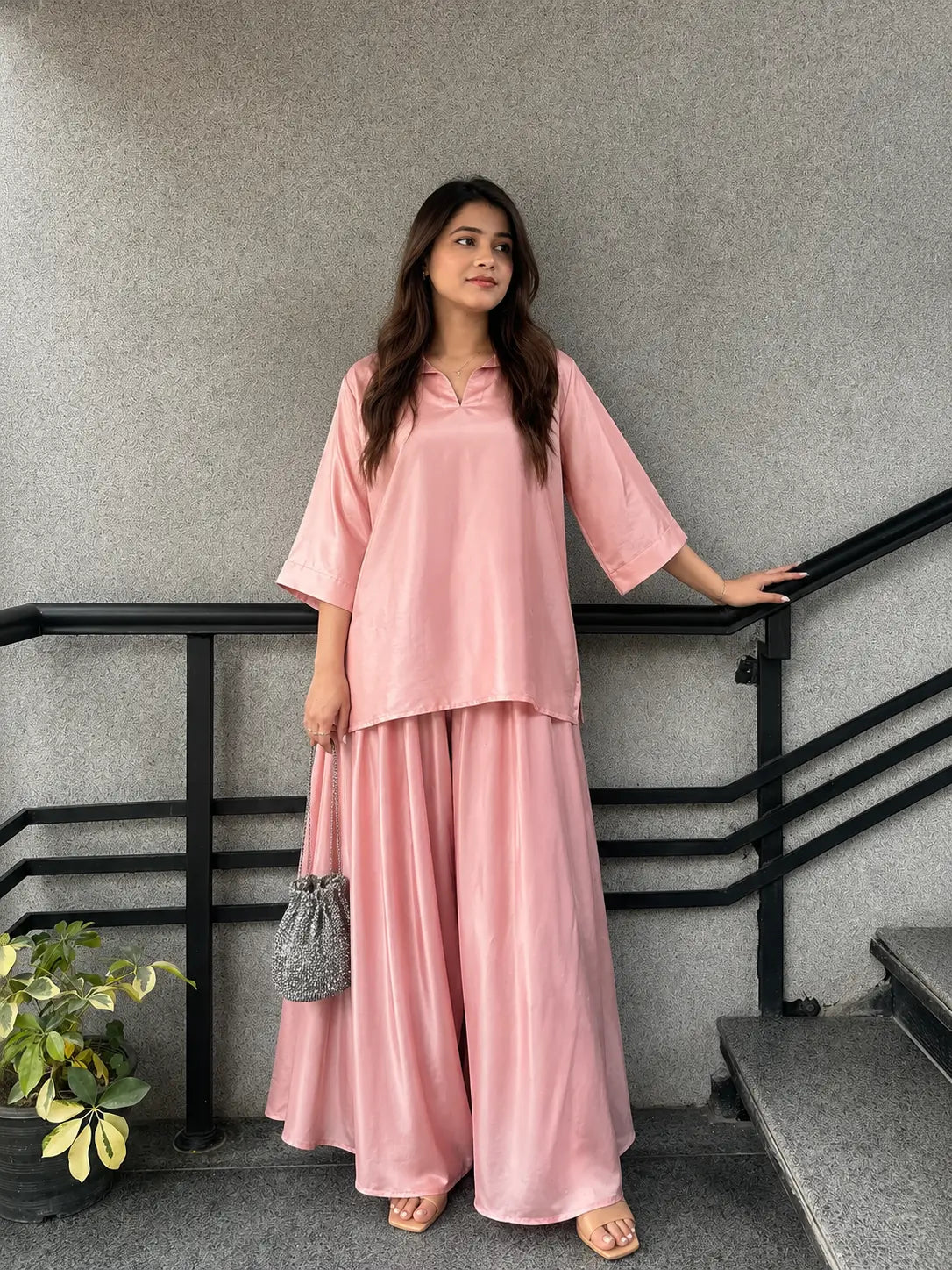 A woman wearing a light pink two-piece outfit consisting of a loose short-sleeve top and matching wide-leg pants, standing on a staircase next to a black railing.